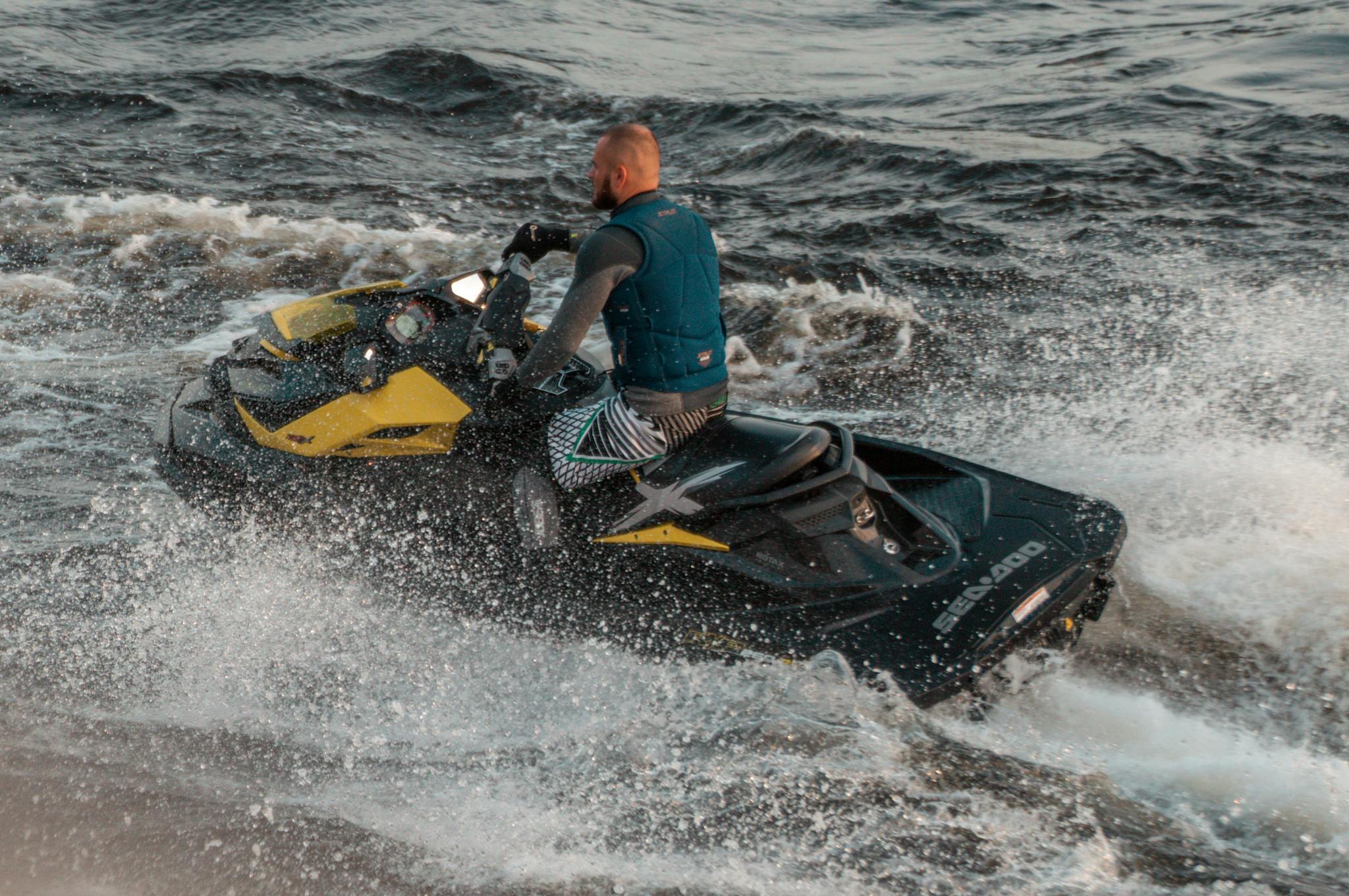 A man wearing a life vest rides a jet ski across choppy waters, creating dramatic splashes.