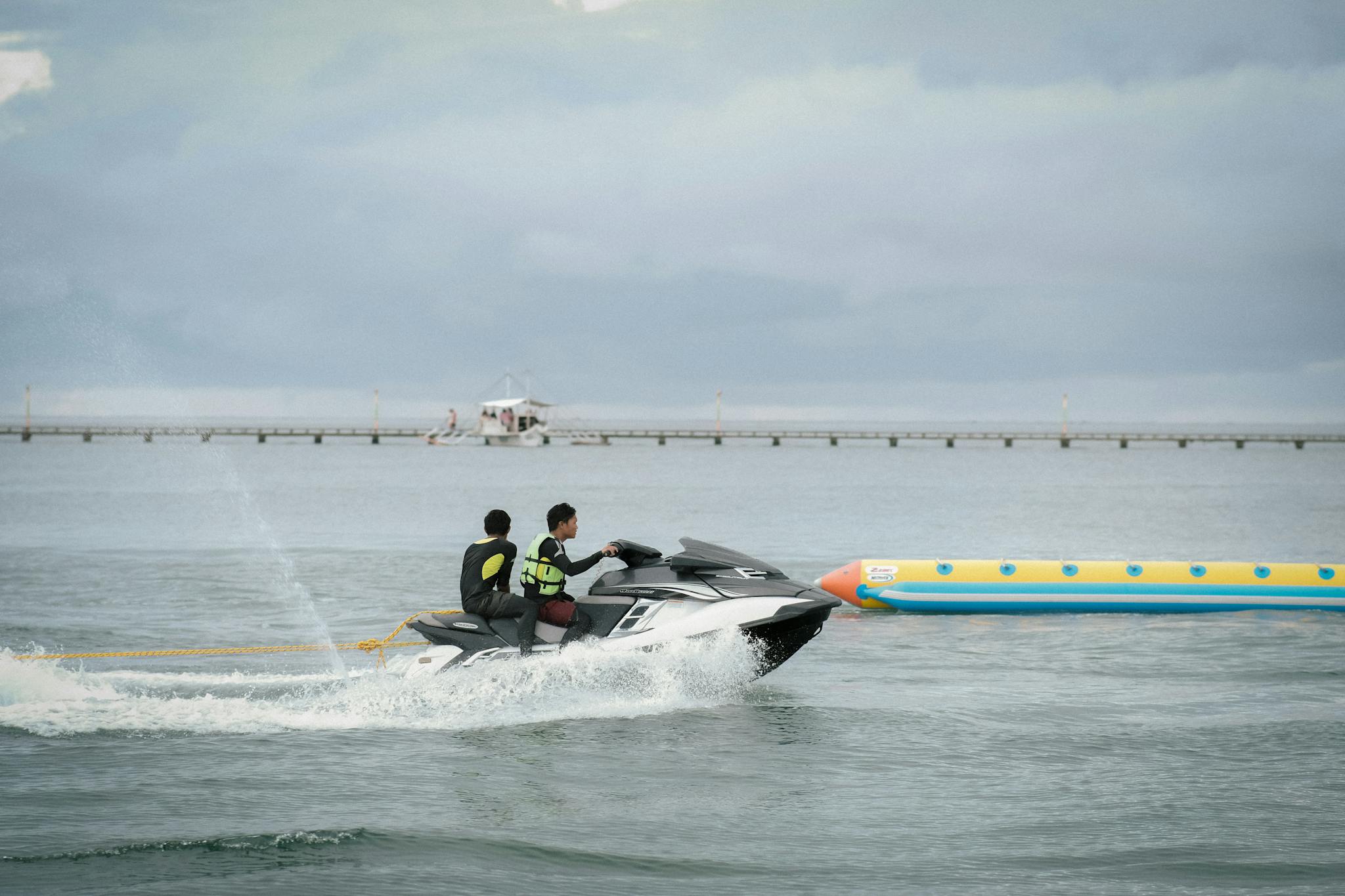 Two people enjoy a thrilling jet ski ride with an inflatable banana boat on a cloudy day.